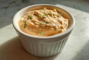 A close-up of thick, creamy, orange-hued remoulade sauce speckled with green herbs and chopped celery.
