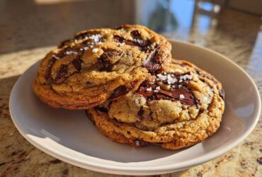 Two thick, golden brown butter chocolate chip cookies topped with flaky sea salt on a white plate.