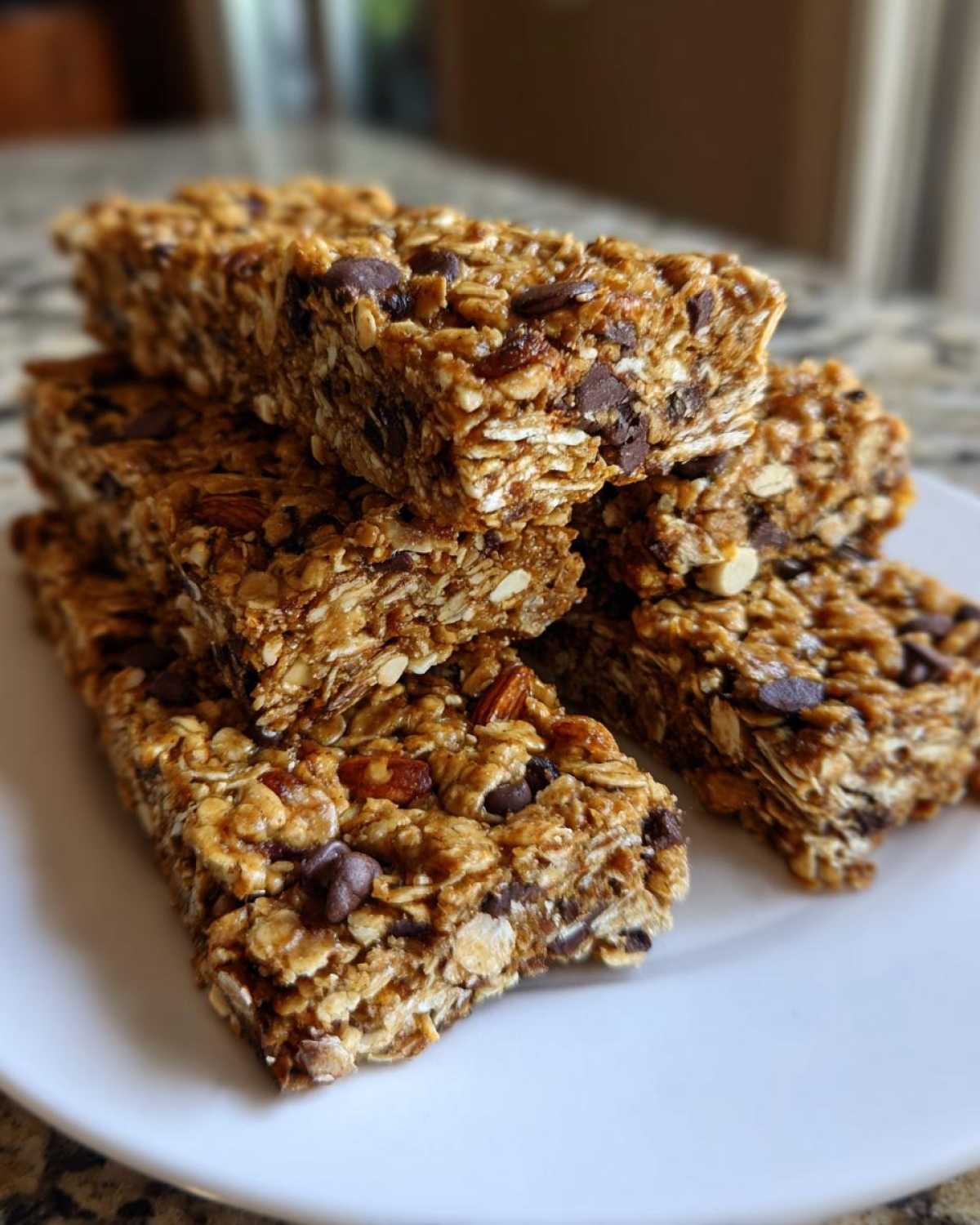 A stack of chewy homemade granola bars featuring oats, nuts, and visible chocolate chips, resting on a white plate.