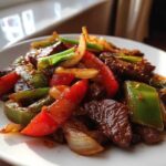 A close-up shot of glistening Chinese pepper steak featuring tender beef strips mixed with bright red and green bell peppers and onions.