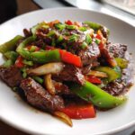 A close-up of glossy Chinese pepper steak mixed with bright green and red bell peppers and onions, garnished with scallions.