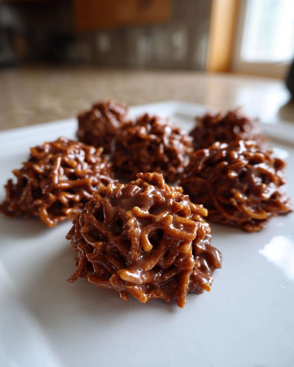Close-up of four glossy, chocolate-coated haystack cookies resting on a white plate.