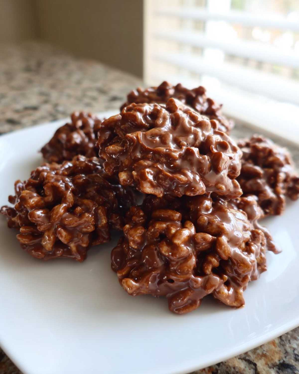 A close-up of several glossy, chocolate haystack cookies piled on a white square plate.