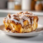 Close-up of a single, gooey cinnamon roll apple pie swirl topped with thick white icing on a small white plate.