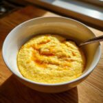 Close-up of a white bowl filled with creamy, yellow cheese grits, topped with melted butter and black pepper.