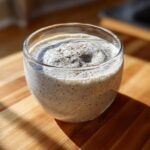 A glass of creamy coconut chia pudding topped with a sprinkle of dark spice on a wooden surface.