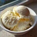 Close-up of creamy, vanilla-colored cottage cheese ice cream scoops served in a white bowl near a window.