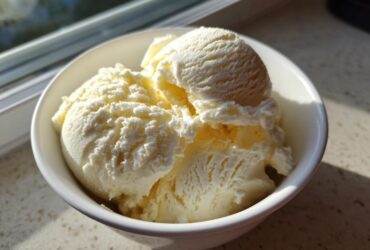 Close-up of creamy, vanilla-colored cottage cheese ice cream scoops served in a white bowl near a window.