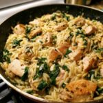 Close-up of creamy lemon chicken orzo with spinach being cooked in a dark skillet.