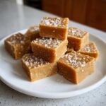 Close-up of creamy maple fudge squares stacked on a white plate, sprinkled with coarse sea salt.