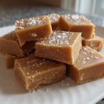 A stack of creamy, light brown maple fudge squares sprinkled with coarse sea salt on a white plate.