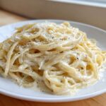 A close-up of creamy parmesan pasta tossed in a rich white sauce and topped with grated cheese and black pepper.