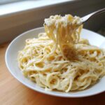 A fork lifting a twirl of creamy parmesan pasta from a white bowl, topped with grated cheese.