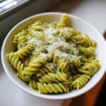 Close-up of a white bowl filled with creamy pesto pasta (fusilli) topped generously with grated Parmesan cheese.