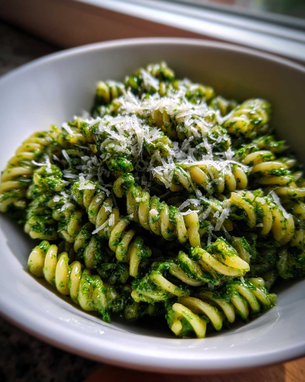 A close-up shot of fusilli pasta tossed in vibrant green creamy pesto pasta, topped generously with grated Parmesan cheese.