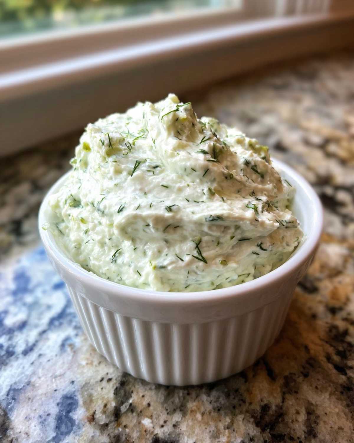 A close-up of creamy, light green pickle dip topped with fresh dill, served in a small white fluted bowl.