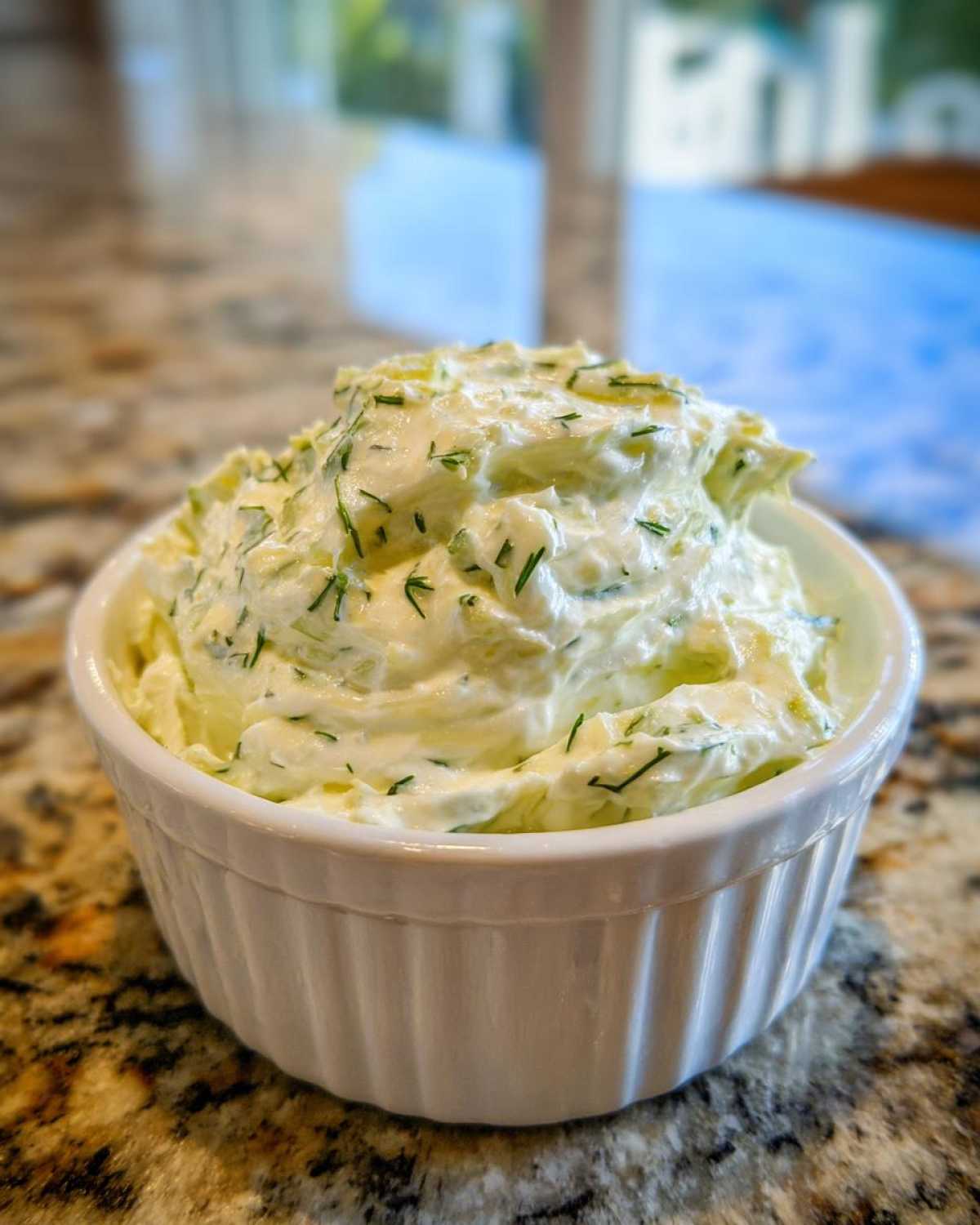 A close-up of creamy, herbed pickle dip served in a small white ramekin on a granite countertop.