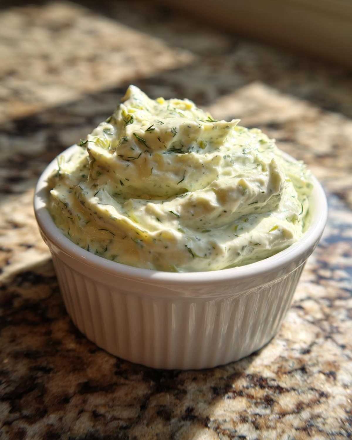 A close-up of creamy, green-flecked pickle dip swirled high in a white ramekin on a granite counter.