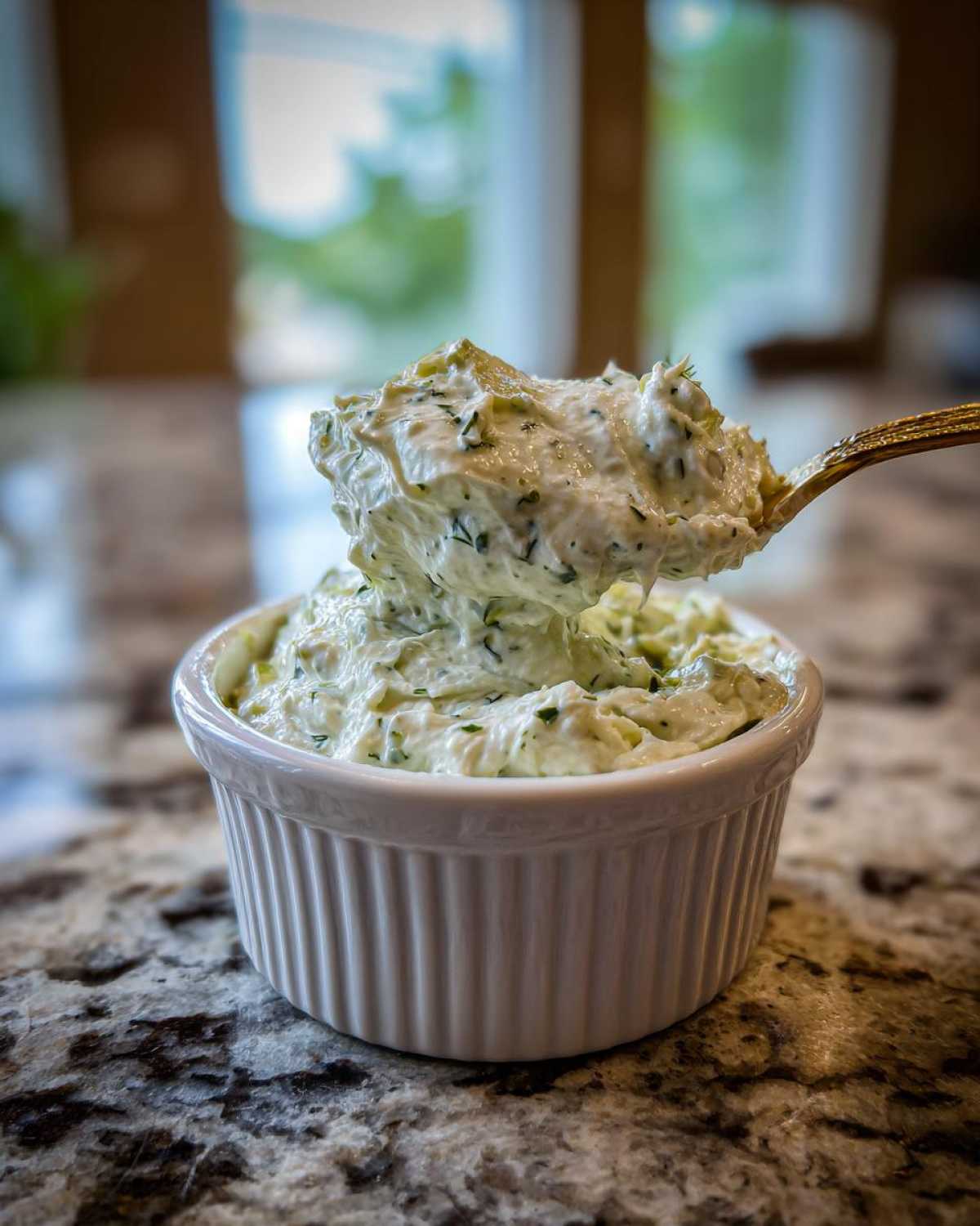 A spoonful of creamy, herbed pickle dip being lifted from a white ramekin on a granite countertop.
