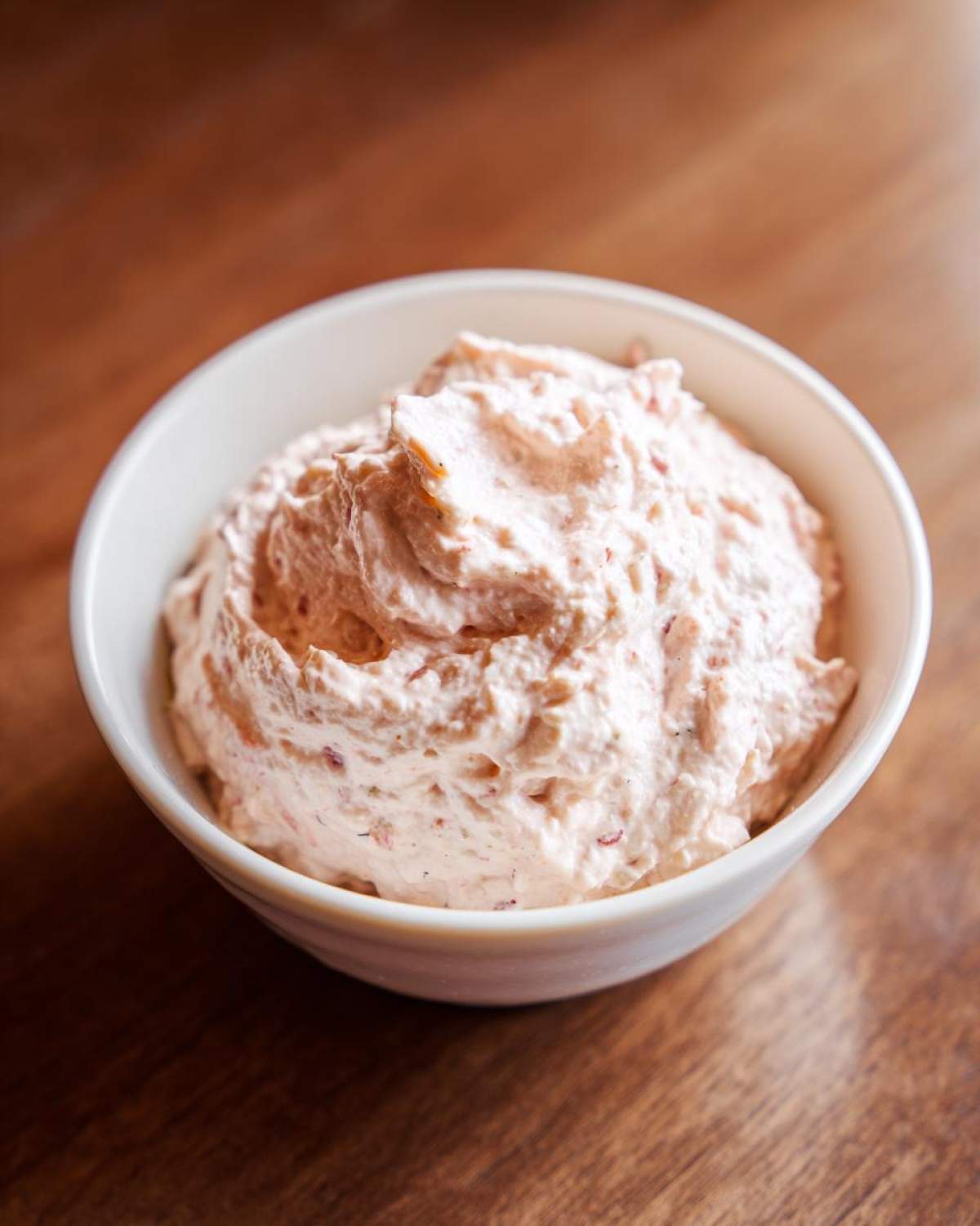 A close-up of a creamy, pinkish tuna dip swirled high in a small white bowl on a wooden surface.
