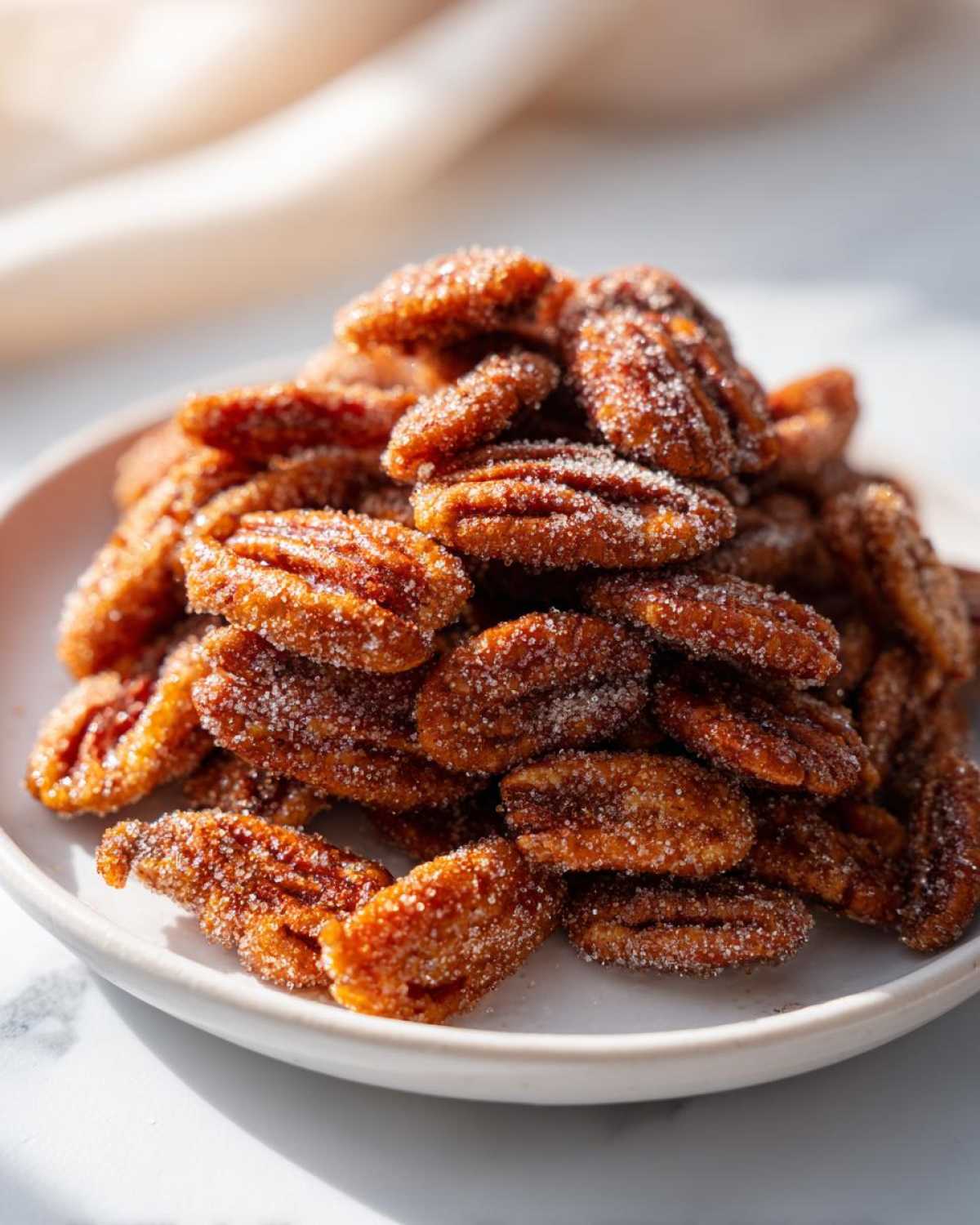A close-up of a mound of freshly made, glistening cinnamon sugar pecans piled high on a white plate.