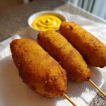 Three freshly fried, golden brown corn dogs on sticks resting on a white napkin, with a small bowl of mustard in the background.