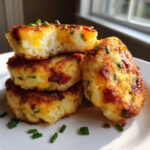 A stack of four golden-brown, crispy mashed potato cakes garnished with fresh chives, one on top is broken open.