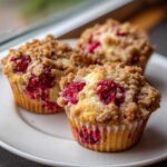 Three freshly baked raspberry muffins with a thick, golden crumb topping, sitting on a white plate.