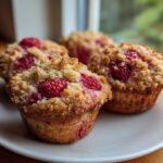 Four golden brown raspberry muffins with a crunchy streusel topping served on a white plate.