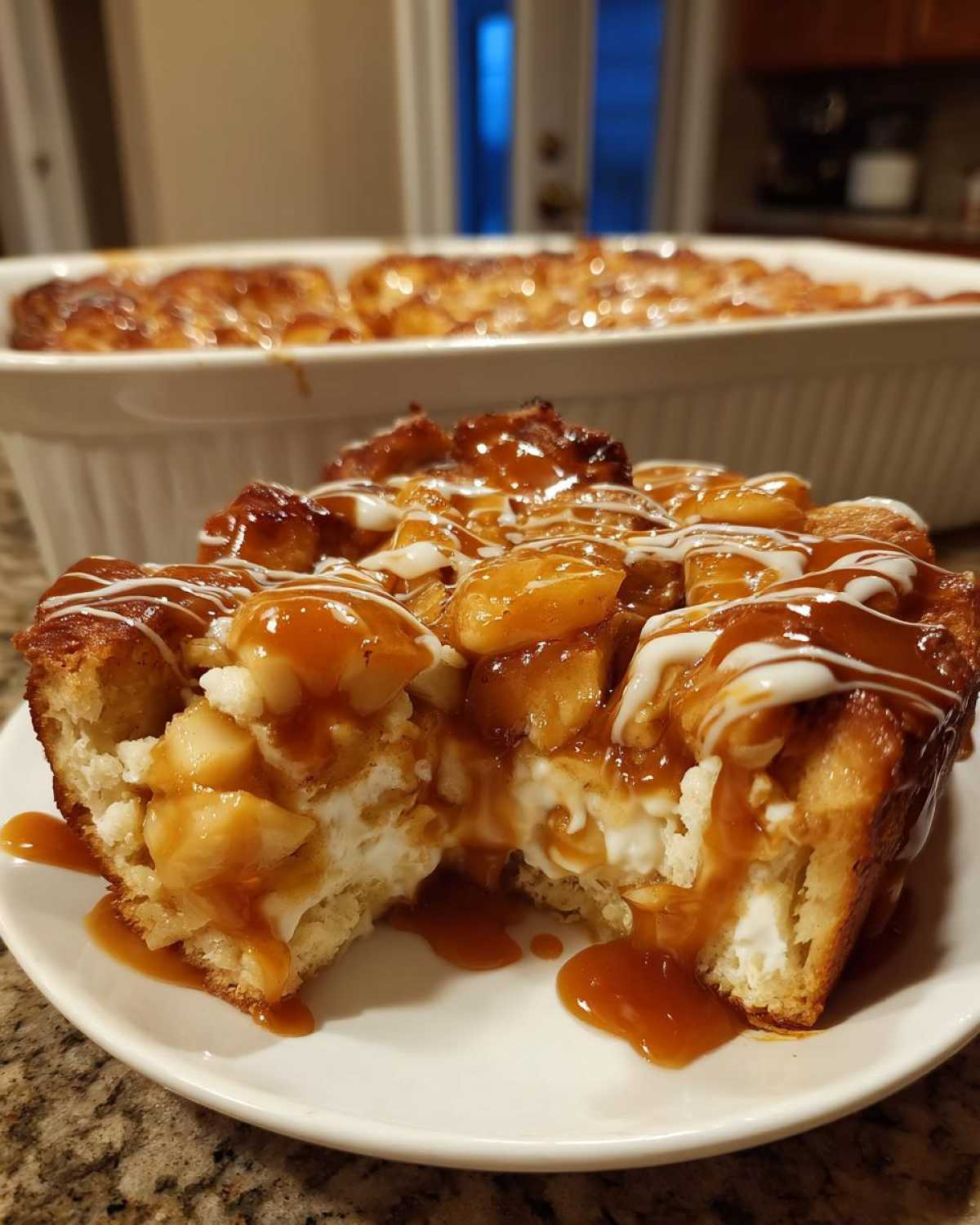 A close-up of a serving of rich apple bread pudding topped with caramelized apples, caramel sauce, and white icing.