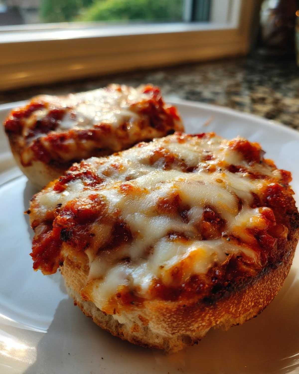 Close-up of two golden brown english muffin pizza halves topped with melted mozzarella cheese and tomato sauce.