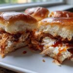 A close-up of one of the bbq chicken sliders cut in half, showing shredded chicken, melted cheese, and BBQ sauce on a shiny brioche bun.
