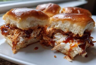 A close-up of one of the bbq chicken sliders cut in half, showing shredded chicken, melted cheese, and BBQ sauce on a shiny brioche bun.