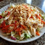 A vibrant plate of thai chicken salad featuring shredded chicken, carrots, cabbage, and topped generously with chopped peanuts.