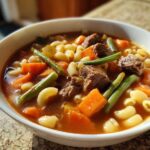 Close-up of a white bowl filled with rich vegetable beef soup featuring chunks of beef, carrots, green beans, and elbow macaroni.