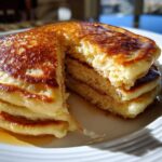 Close-up of a fluffy stack of banana pancakes with a piece cut out, drizzled generously with syrup.
