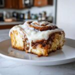 A close-up of a single, fluffy cinnamon bun topped generously with dripping white frosting.