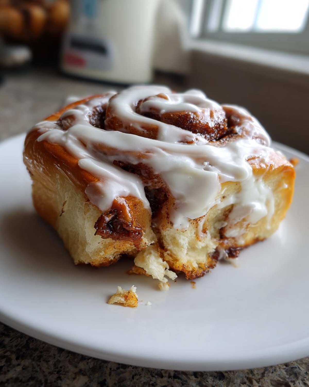 A close-up of a fluffy, freshly baked cinnamon bun topped with thick white icing, sitting on a white plate.