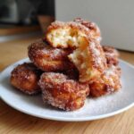 A stack of golden brown, sugar-coated buñuelos, with one broken open to show the fluffy interior.