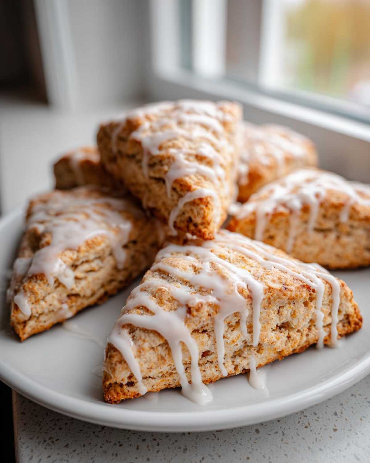 Close-up of several fluffy lemon scones drizzled generously with a bright white, tangy glaze, stacked on a white plate.