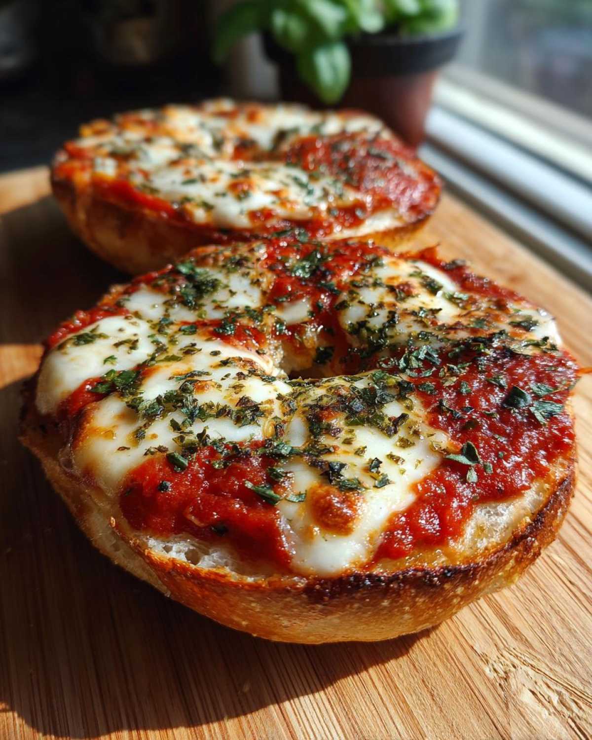 Two freshly baked pizza bagels topped with bright red sauce, melted mozzarella cheese, and dried herbs, sitting on a wooden board.