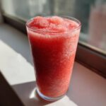 A tall glass filled with a bright red, icy frozen strawberry daiquiri, sitting on a sunlit windowsill.