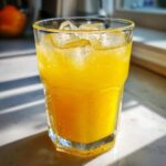 Close-up of a tall, sweating glass filled with bright yellow homemade lemonade and ice cubes, sitting in sunlight.