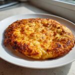 A perfectly baked, golden brown keto pizza crust with a bubbly, cheesy top sitting on a white plate.