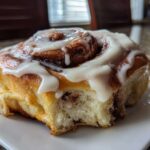 A close-up of a fluffy, freshly baked cinnamon bun generously topped with thick white icing.