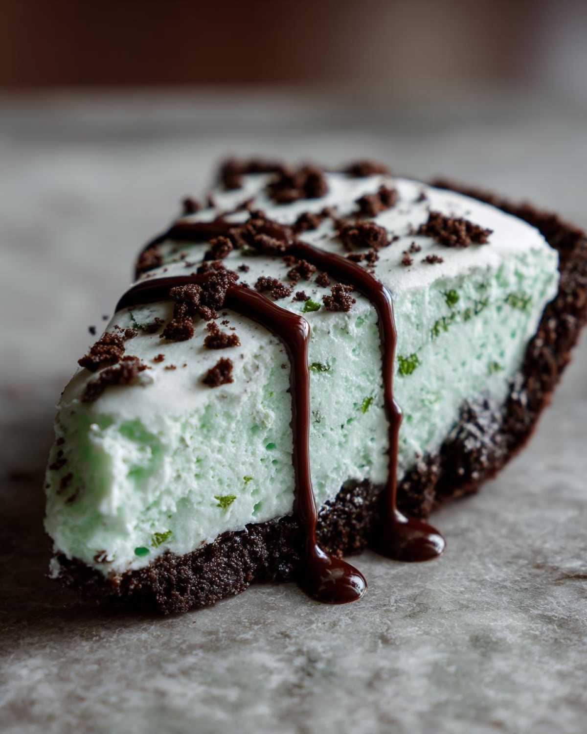 A close-up of a slice of light green grasshopper pie with a dark chocolate crust, topped with chocolate syrup and crumbs.