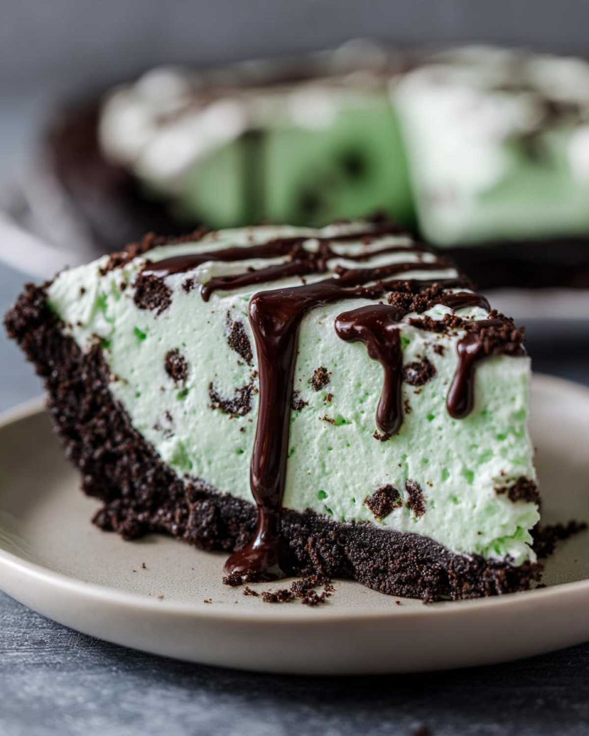 A close-up slice of mint green grasshopper pie with a dark chocolate cookie crust, topped with chocolate syrup.