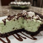 A close-up slice of mint green grasshopper pie with a dark chocolate crust, topped with marshmallows and chocolate syrup.