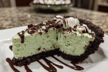 A close-up slice of mint green grasshopper pie with a dark chocolate crust, topped with marshmallows and chocolate syrup.