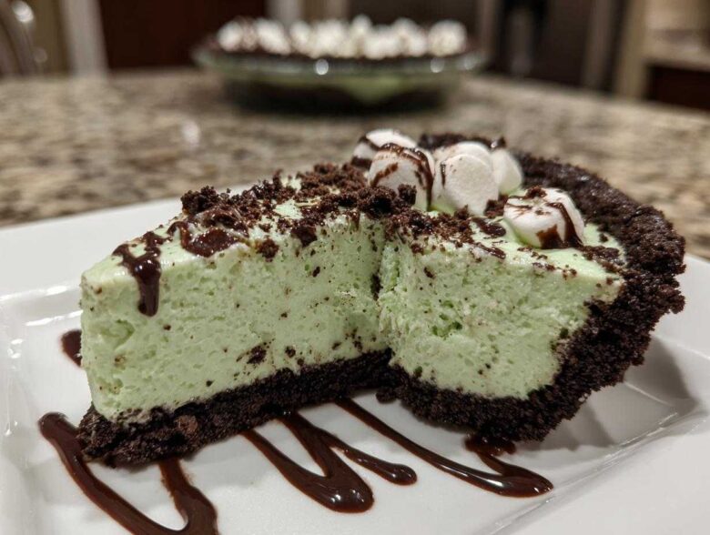 A close-up slice of mint green grasshopper pie with a dark chocolate crust, topped with marshmallows and chocolate syrup.